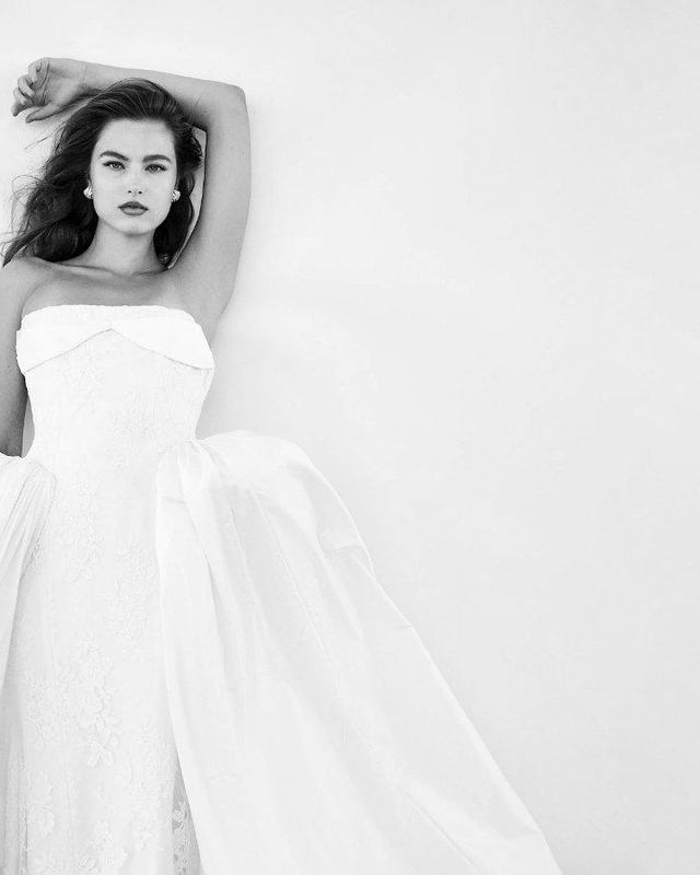A bride in a strapless Houghton gown with one arm raised against a white wall